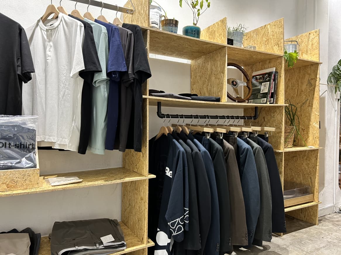 木目調のオープン棚にTシャツやジャケットが整然と並び、ハンガーラックに衣類が掛けられたシンプルで落ち着いた雰囲気の店内